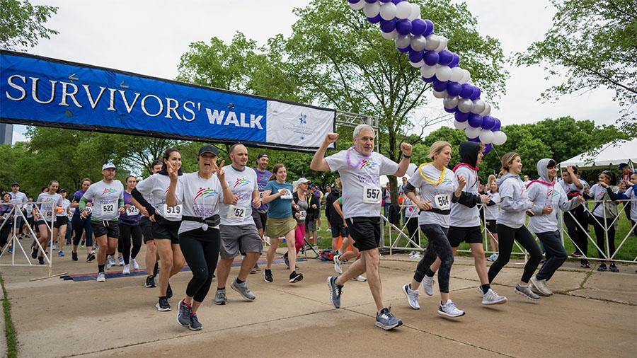 A crowd of runners start racing along the lakeshore path under a purple survivors' banner.