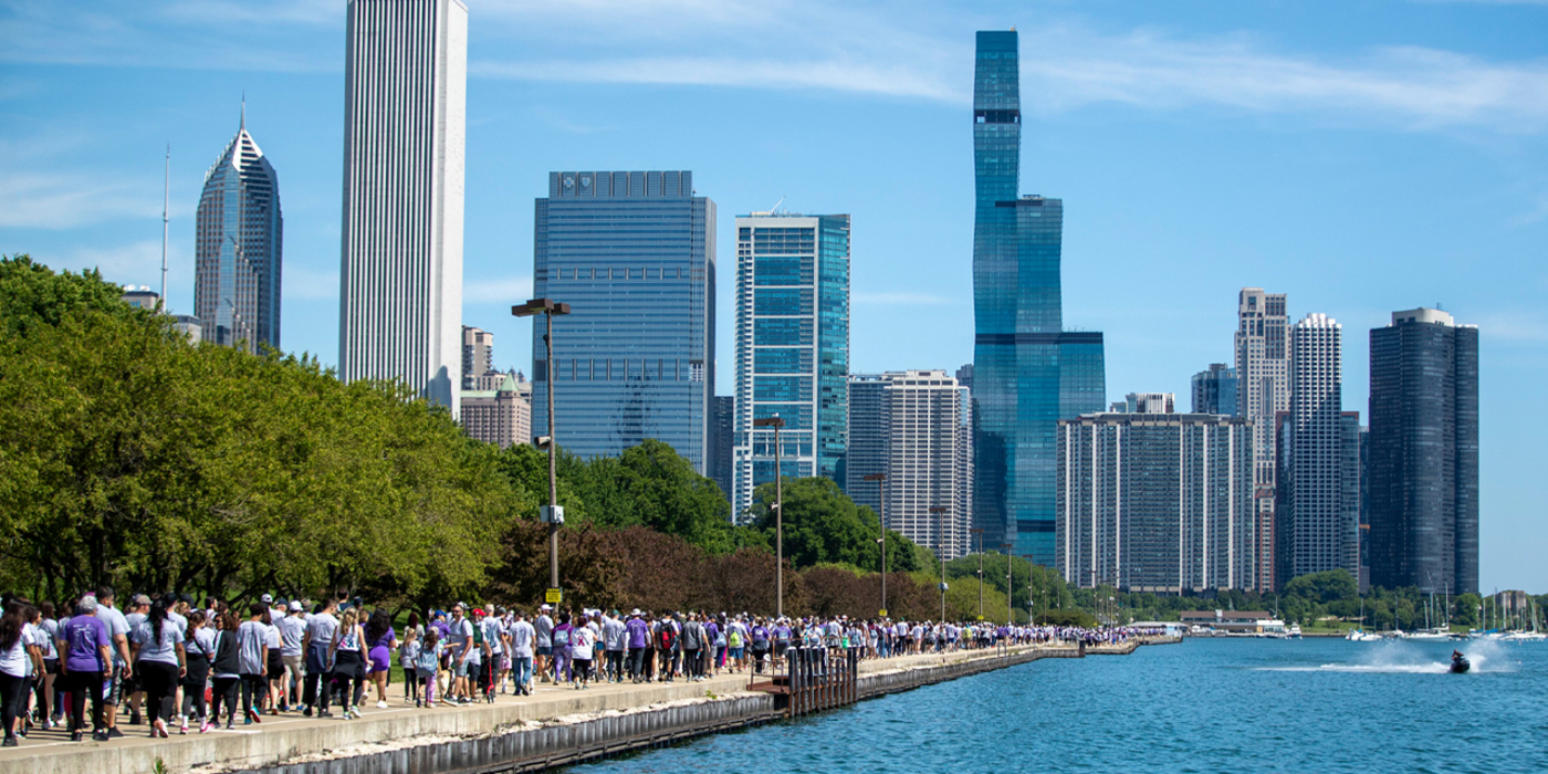 Large crowd of participants walks along the Lake Michigan shoreline with Chicago skyline in background.